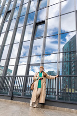 A 60-year-old woman, wrapped in a beige trench, walks amidst the towering structures. Her presence adds a touch of grace to the bustling metropolis.