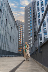 Elegant Caucasian Woman in Beige Coat Strolling Amidst Urban Skyscrapers and Smiling. Urban style. Concept of beautiful old age and active longevity.