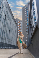 A 60-year-old woman in a beige raincoat walks among tall buildings. Full length view. Urban style. Concept of beautiful old age and active longevity.