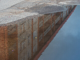High-rise buildings with red columns reflected in a rain puddle on textured asphalt. The surface has grainy details and a visible space for text.