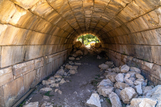 Close-up of an ancient vaulted vomitorium at Patara Theater, partially collapsed and filled with debris. Sunlight illuminates green vegetation through the archway. Patara, Mediterranean, Turkey.

