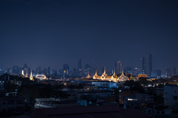 Fototapeta premium Nighttime view of the Grand Palace illuminated against the modern Bangkok city skyline, with skyscrapers form the backdrop of Thailand's capital city.