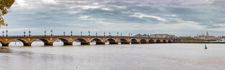 Fototapeta premium Panoramic view of Pont de Pierre, a stone bridge with multiple arches and lampposts, spanning the Garonne River under a cloudy sky. Bordeaux, Gironde, Nouvelle-Aquitaine, France.