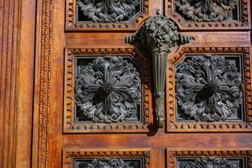 An intricate wooden door features ornate carvings, decorative metal rosettes, and a large horn-shaped bronze handle.