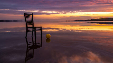 A surreal photograph of a black wooden chair standing in shallow water at sunset.