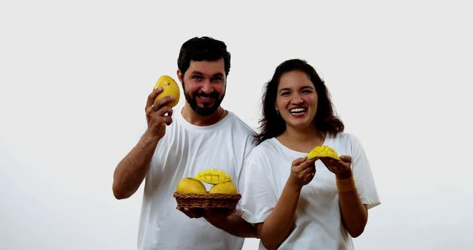 Indian young couple eating mango while standing isolated against white background, holding, promoting fresh seasonal aam or Alphonso happily, enjoying king of fruits with joyful expressions smiles