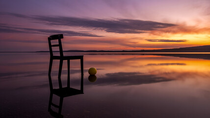 A surreal photograph of a black wooden chair standing in shallow water at sunset.