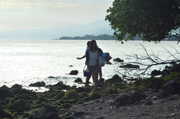 Teenagers step slowly across moss-covered coral near a beachside tree, cautiously carrying tent and...
