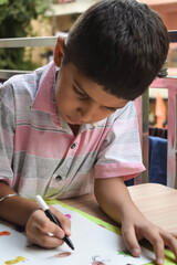 Smart Indian little boy perform thumb painting with different colourful water colour kit during the summer vacations, Cute Indian Kid doing colourful thumb painting drawing on wooden table