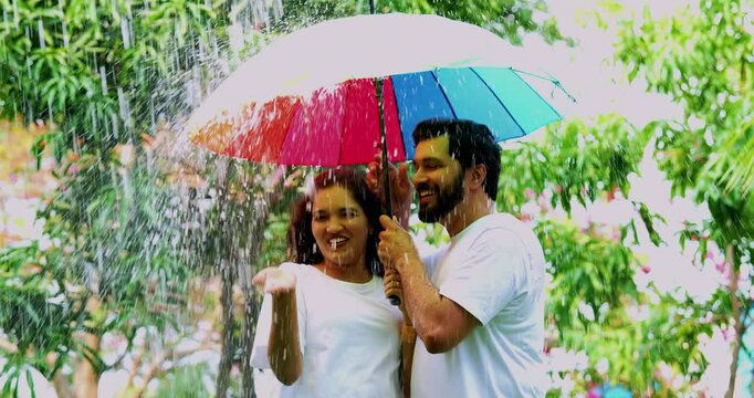 Couple enjoying rain under umbrella, Indian Asian young man woman standing outdoors in park during first monsoon shower, smiling, playing with raindrops, holding colorful umbrella happily together