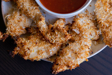  chicken fillets coated with coconut shreds, placed on a plate with sweet chili sauce. Tropical flavor dish, crispy texture, rich taste 