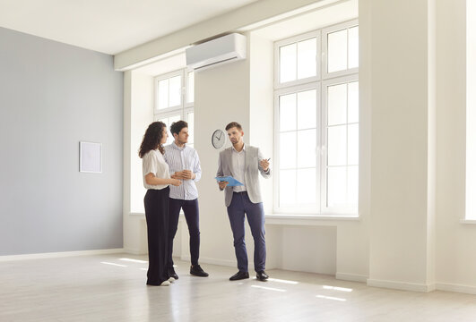 Professional male realtor showing empty apartment to young couple during real estate tour. Confident agent presenting advantages of modern flat and consulting clients about terms of purchase or rent. - Powered by Adobe