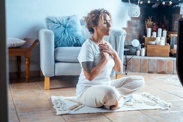 Mature curly-haired woman practicing yoga and mindfulness at home. Middle-aged female sitting on floor in meditation or wellness pose. Concept of healthy lifestyle, relaxation, balance and self care