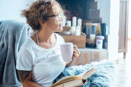 Beautiful mature woman relaxing at home in peaceful atmosphere. Middle-aged curly-haired female enjoying quiet time indoors, reading a book and drinking herbal tea. Concept of comfort, wellbeing, slow