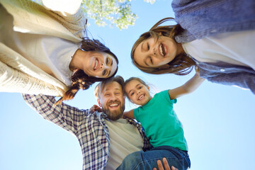 Bottom view of happy young family of four looking cheerful at camera outdoors on blue sky background. Portrait of smiling mother, father and children enjoying time in nature together.