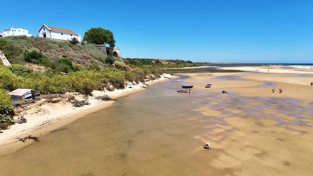Aerial view of Cacela Velha, Ria Formosa, Algarve, in summer, capturing crystal-clear waters, golden sand, traditional boats, and people enjoying the beach.