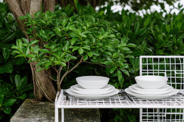 Minimalist table setting with white ceramic dishes arranged on modern wire rack, surrounded by lush green foliage, creating serene outdoor dining atmosphere