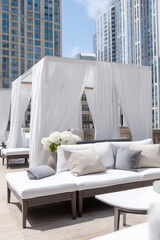 A luxurious rooftop dining area featuring oversized white sofas and elegant drapes, surrounded by modern cityscape buildings and adorned with fresh white flowers