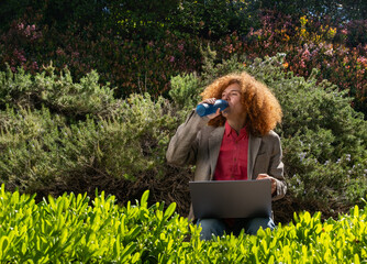 Young man with curly hair drinking from a reusable bottle while working on a laptop outdoors, surrounded by greenery. Concept of sustainable remote work and eco-friendly lifestyle.