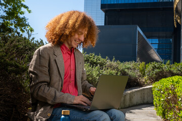 Young man with curly hair smiling while working on a laptop outdoors, reusable bottle in foreground. Concept of happy remote work, eco-friendly business and sustainable digital lifestyle.