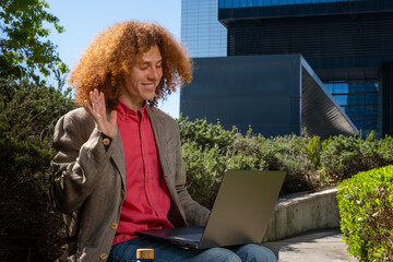 Young man with curly hair waving during a video call outdoors, working on a laptop with reusable bottle visible. Concept of remote communication, sustainable digital work and eco-friendly lifestyle.