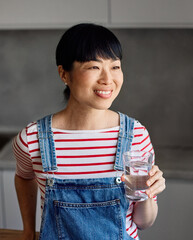Portrait of a young happy asian woman drinking water and standing in the kitchen
