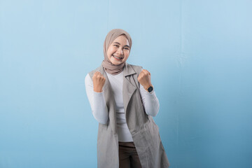 A woman in a grey vest is smiling and raising her arms. She is wearing a head scarf and she is happy