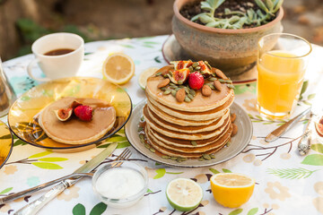 Stack of pancakes with fresh fig, strawberries, maple syrup, and crunchy toppings. A warm, comforting outdoor breakfast with orange juice and coffee