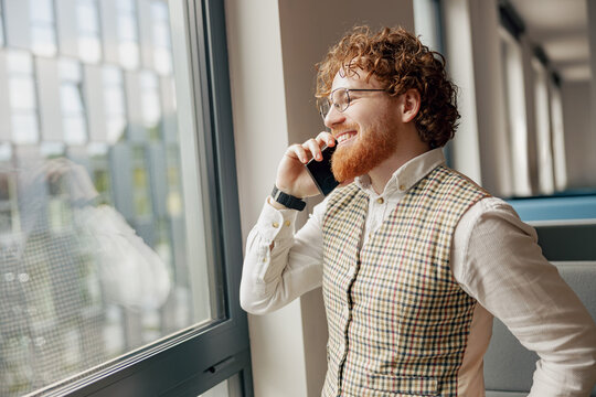 A young man in a stylish vest is confidently taking a phone call by a large window in a bright modern office