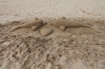 Two dolphins sculpted from sand lie nose-to-nose on a beach with a heart shape between them, creating a playful and romantic seaside scene full of charm and creativity.