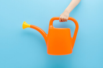 Obraz premium Little child boy hand holding and showing orange plastic can for flowers and plants watering on light blue wall background. Pastel color. Closeup. Front view.
