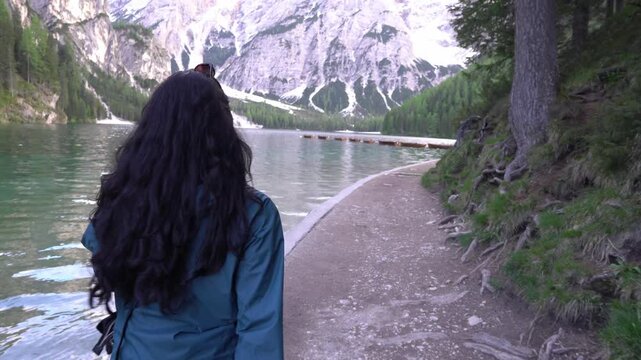 A woman in black pants and a turquoise rain jacket walks along the scenic lakeside trail at Lago di Braies in the Italian Dolomites