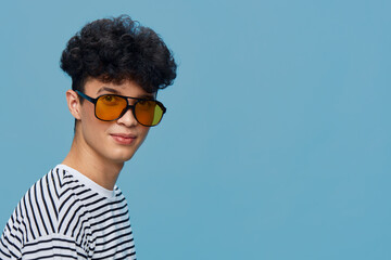 Young man wearing yellow sunglasses and striped shirt stands against blue background with curly...
