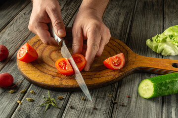 Fresh tomatoes are being sliced on a wooden cutting board, accompanied by a cucumber and lettuce....