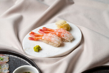 Succulent shrimp sushi rests gracefully on a polished white plate, accompanied by a dab of vibrant wasabi and pickled ginger. A soft, textured fabric creates an inviting atmosphere
