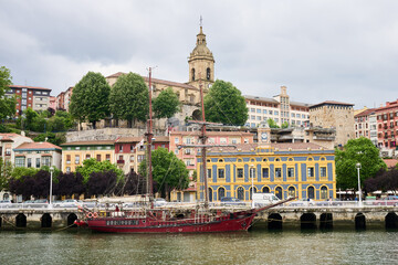 Red sailing ship docked on nervion river, with colorful buildings of portugalete's ribera district...