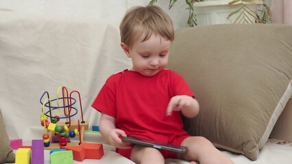 Naklejka premium Adorable toddler boy sitting on the sofa in the living room and playing with smartphone. Child learning how to use smartphone. Boy texting on the phone. - technology and lifestyle concept