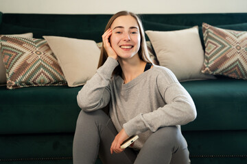 Obraz premium Young woman smiling at camera holding smartphone in hand enjoying leisure time wearing grey outfit sitting on floor in cozy modern home interior, lifestyle indoors, digital technology, positive mood