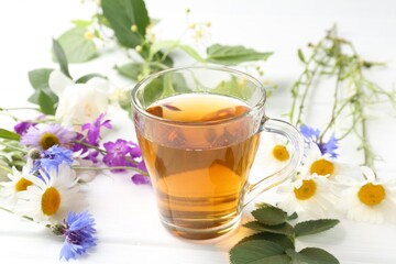 Aromatic herbal tea in glass cup, different flowers and green leaves on white wooden table, closeup