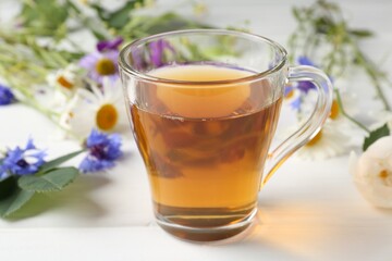 Aromatic herbal tea in glass cup and different flowers on white wooden table, closeup