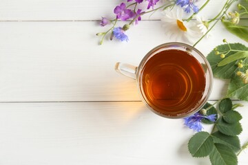 Aromatic herbal tea in glass cup, different flowers and green leaves on white wooden table, flat...