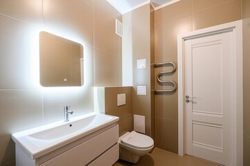 modern bathroom with beige tiles. It features a lighted mirror above a white vanity, a toilet, and a white door. A heated towel rack is mounted on the wall