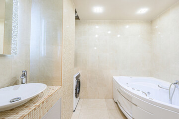 Bright bathroom with beige tiling. It features a modern sink, corner bathtub, and a washing machine in a clean, simple design