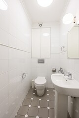 tidy, small bathroom with white tiled walls, a toilet, a sink, and a mirrored cabinet. The floor has a grey and white tiled pattern. Minimalist design
