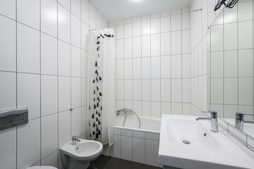 bright bathroom with white tile walls, a bathtub with a shower curtain, a sink, and a bidet. Minimalist and clean design