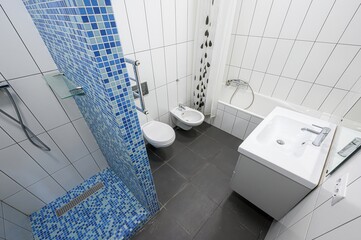 modern bathroom with white tiles, a blue mosaic shower partition, toilet, bidet, bathtub, and sink....