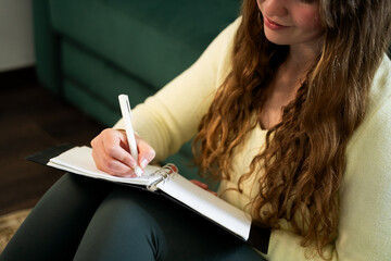 Smiling young woman sitting on floor at home and writing in notebook journaling taking notes in...