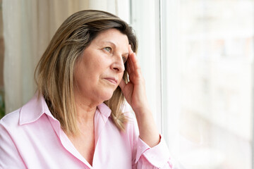 Worried senior woman touching her head in contemplation near a window at home, expressing anxiety...