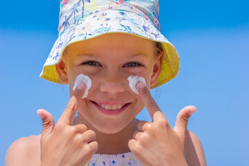 Child happily applies sunscreen on her face, wearing a bright sun hat, with a clear blue sky in the background
