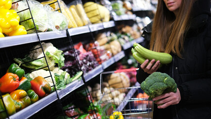 Young caucasian woman choosing zucchini and broccoli in supermarket
